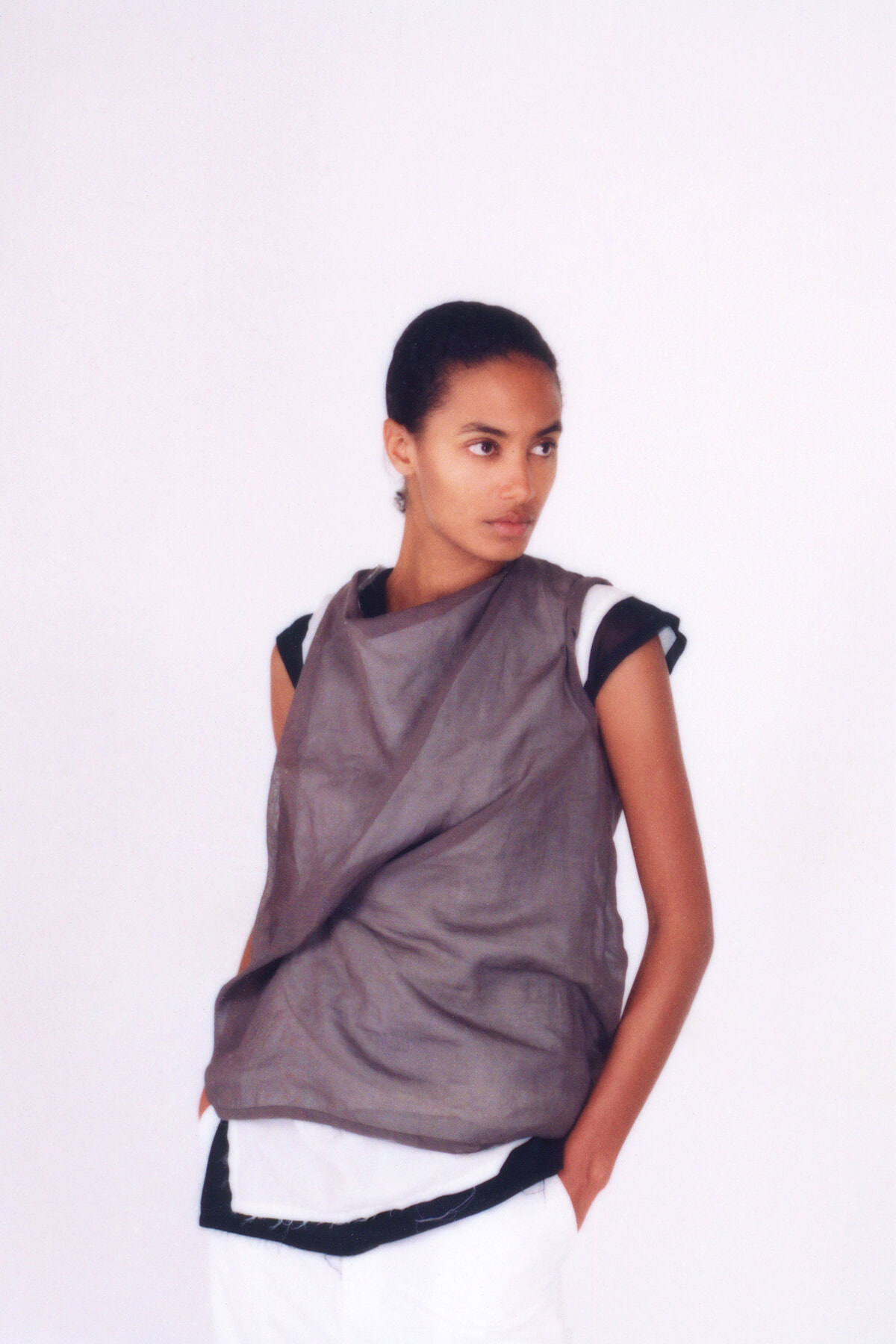 Model wearing a loose taupe draped top over black-and-white layered clothing, standing with hands in pockets against a white backdrop.