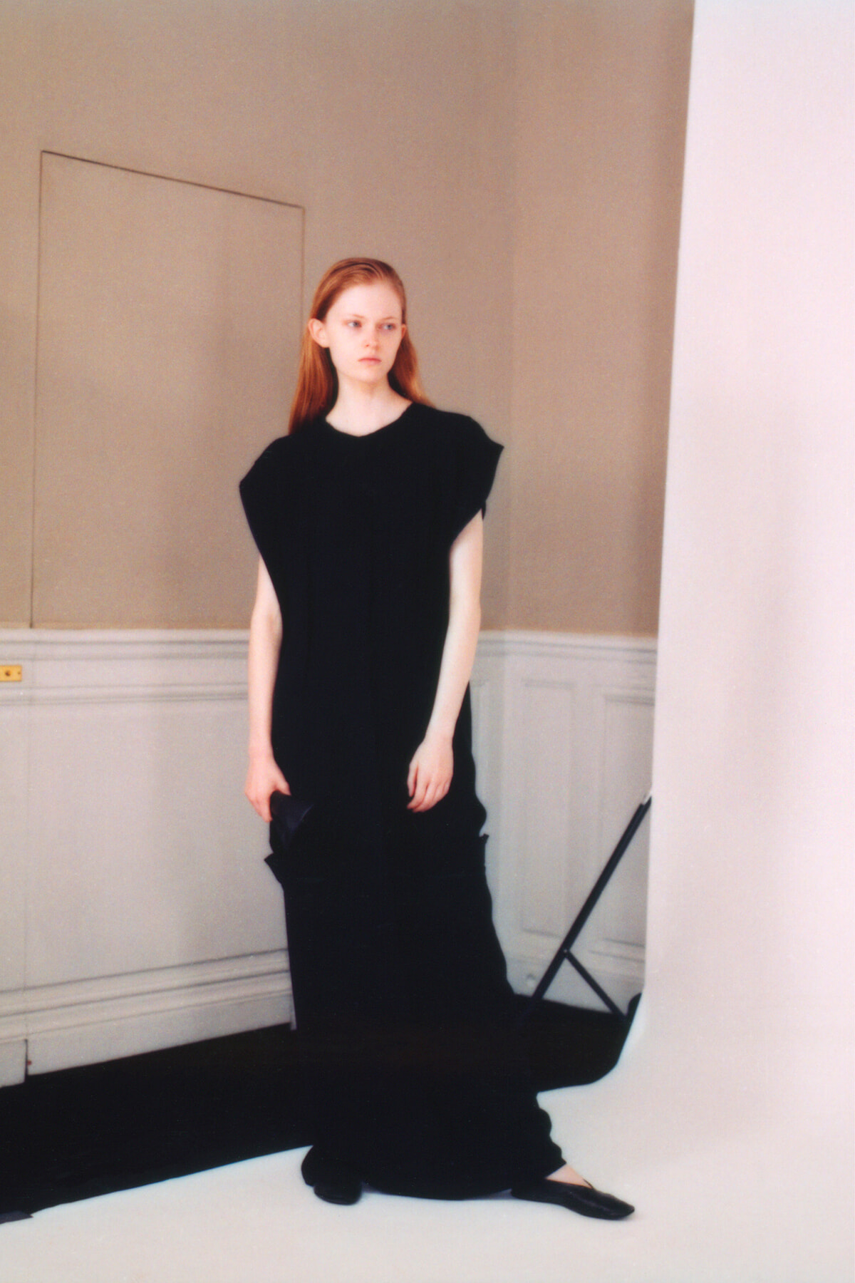 Young woman with red hair in a long black dress standing in a pale studio backdrop.