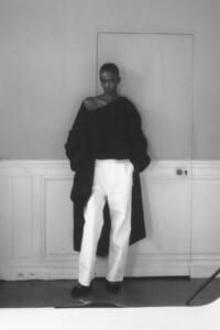 Model stands against a paneled wall, wearing a black oversized coat draped off one shoulder and white wide-leg pants.