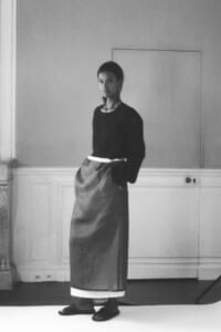Woman stands in a studio wearing a black top and long gray skirt with a white hem, hands in pockets.