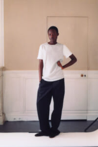 Young person standing in a neutral room, wearing a white short-sleeve top and dark wide-leg pants with one hand on hip.