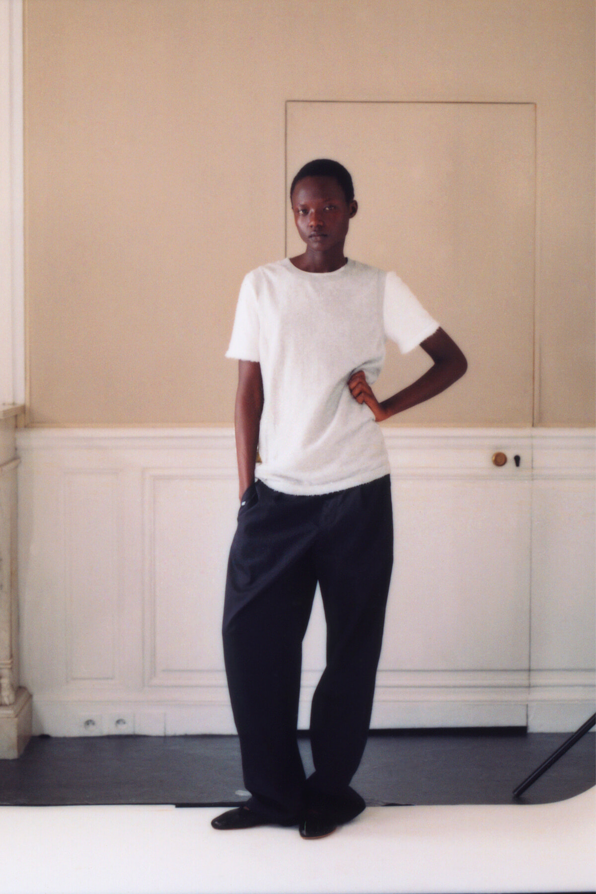 Young person standing in a neutral room, wearing a white short-sleeve top and dark wide-leg pants with one hand on hip.