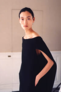 Portrait of a woman with dark hair in a black off-shoulder cape dress, standing in a light room with one hand in her pocket.