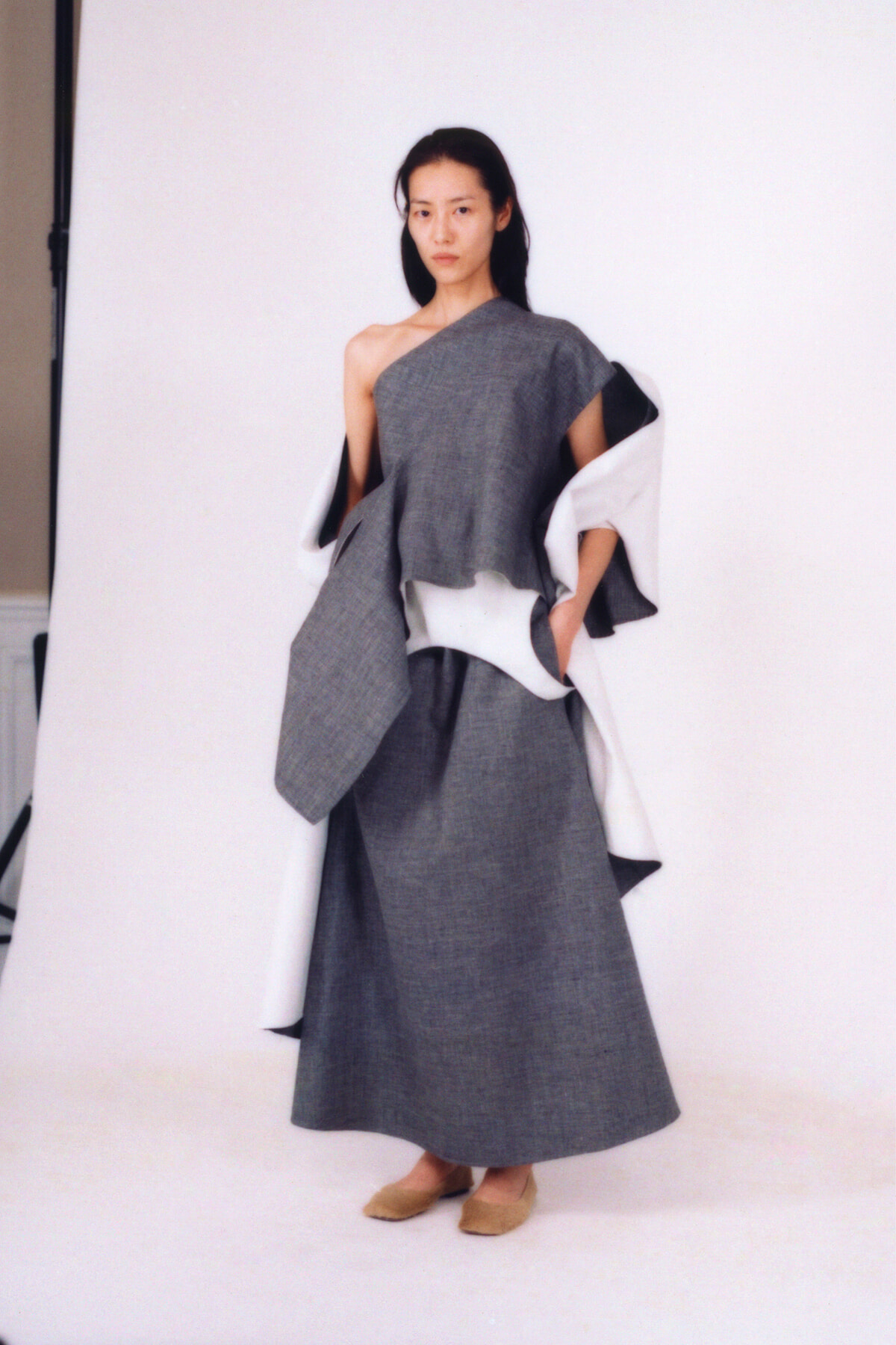 Model in a gray one-shoulder, floor-length dress with a voluminous draped silhouette, paired with a white/gray wrap draped over the arms, standing against a white backdrop in beige flats.