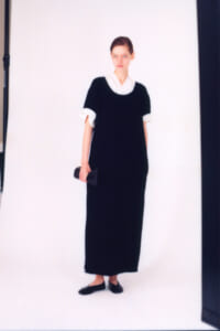 Model in a black ankle- or floor-length dress with a white collar and cuffs, holding a small black clutch in a studio photo.