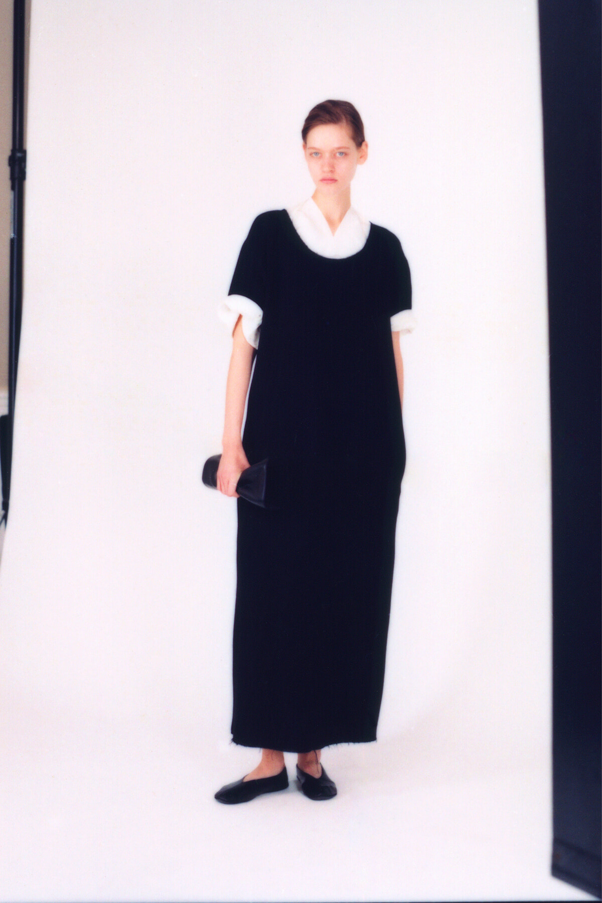 Model in a black ankle- or floor-length dress with a white collar and cuffs, holding a small black clutch in a studio photo.