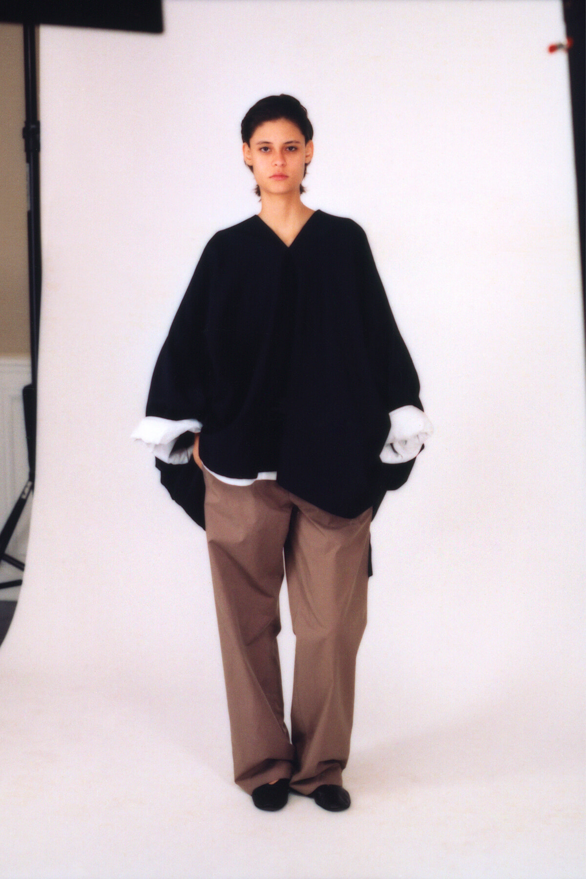 Studio portrait of a person wearing an oversized black blouse with white cuffs, tan wide-leg pants, and black shoes, standing against a white backdrop with a neutral expression.