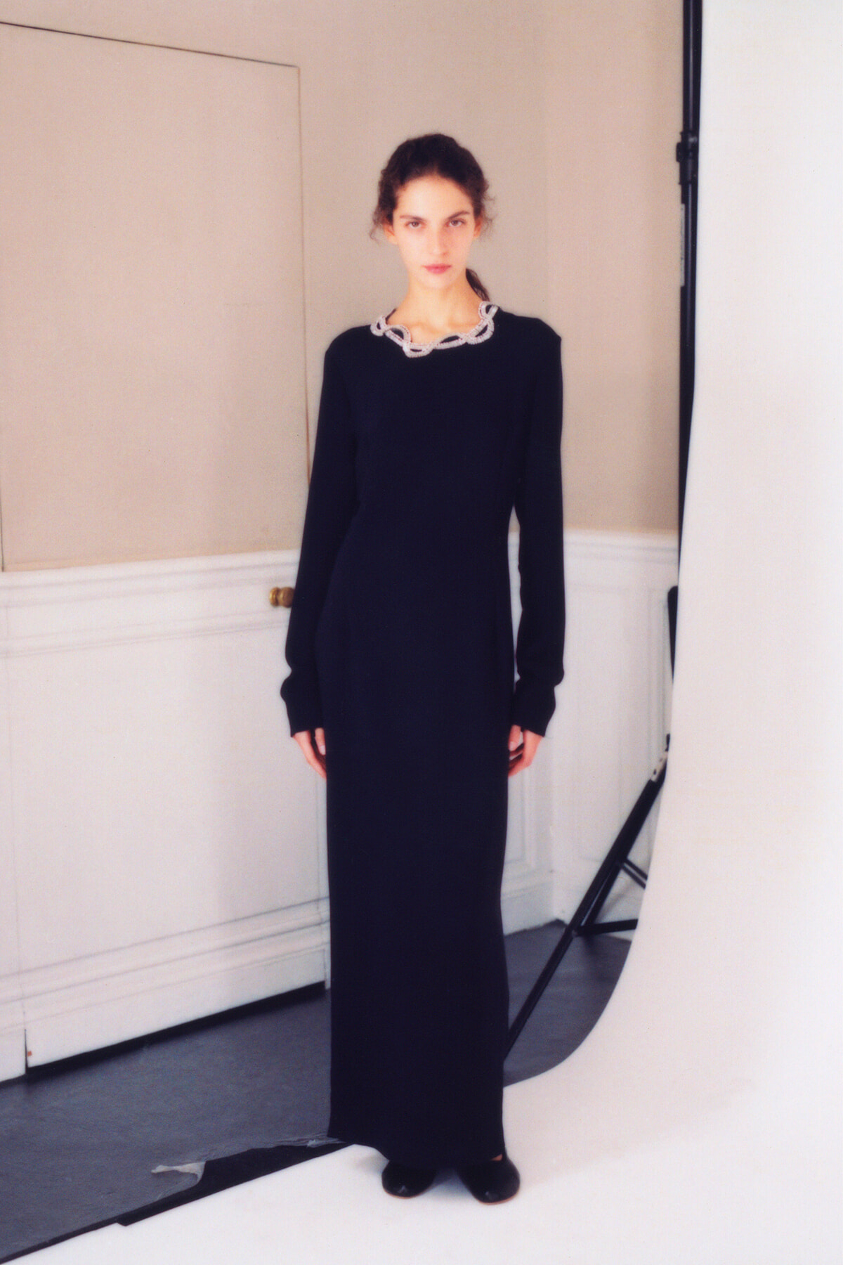 Model wearing a floor-length black dress with a white chain-like neckline, standing in a studio against a light wall.