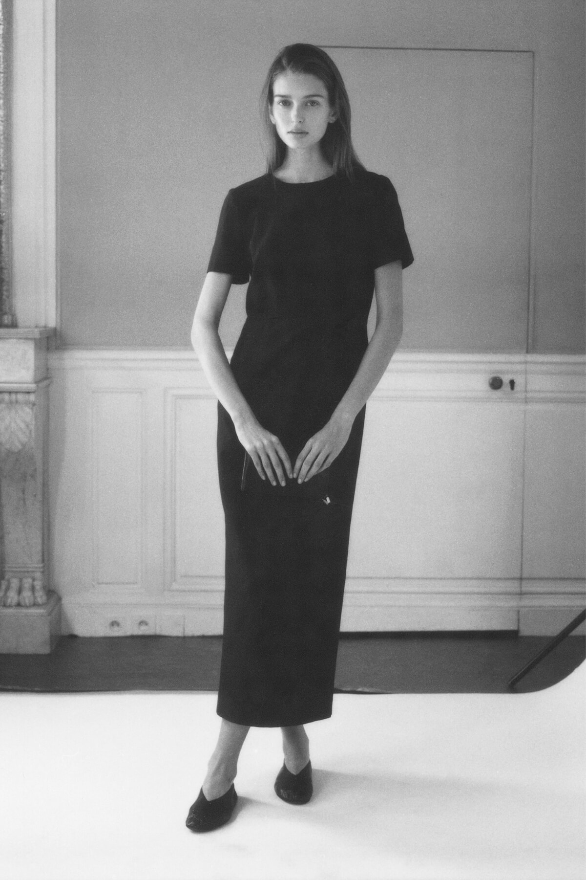 Woman in a black midi dress stands in a photography studio, arms relaxed in front.