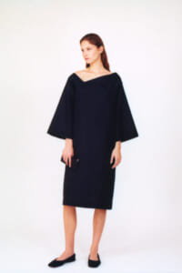 Woman in a dark, off‑the‑shoulder dress with wide, batwing sleeves, standing against a white backdrop and holding a small clutch.