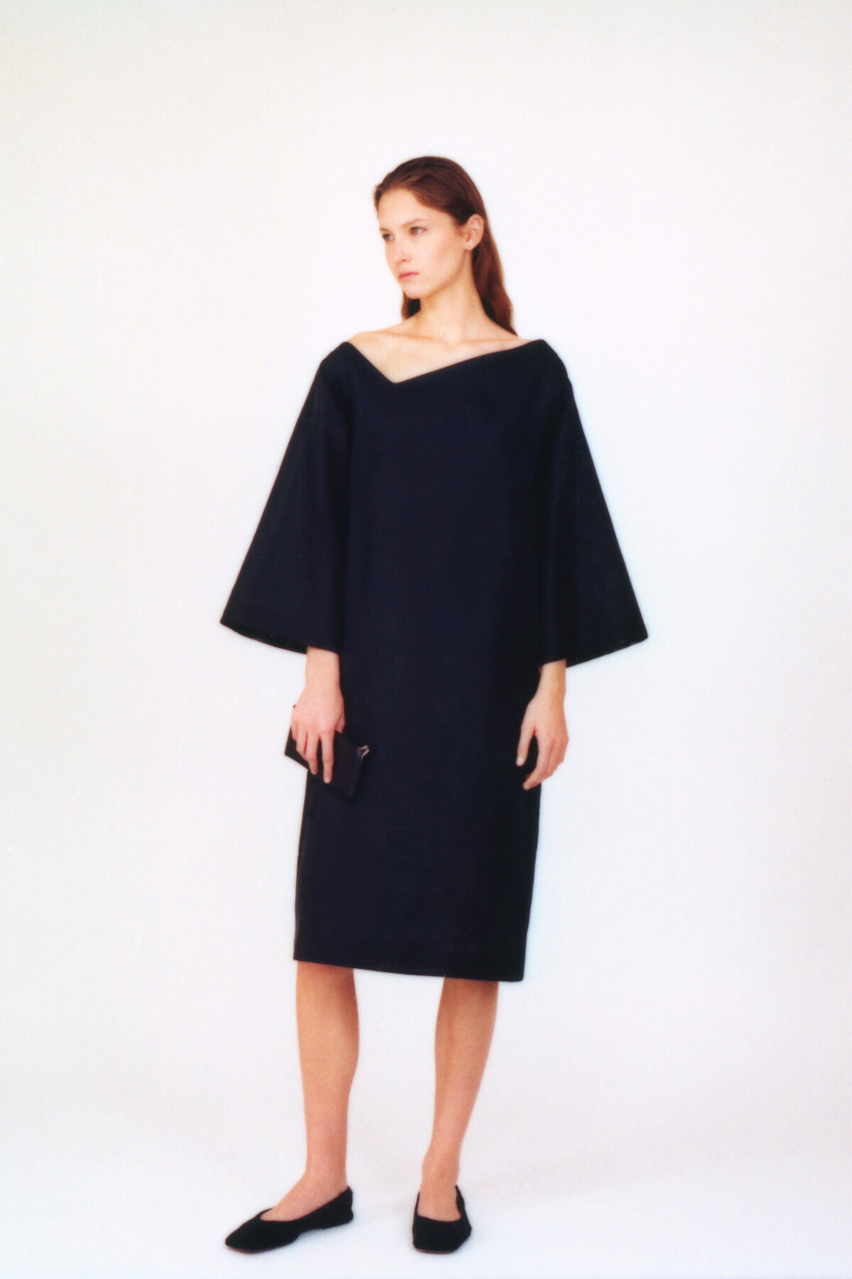 Woman in a dark, off‑the‑shoulder dress with wide, batwing sleeves, standing against a white backdrop and holding a small clutch.