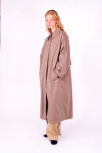 Full-body studio portrait of a woman wearing a long beige trench coat with hands in pockets, neutral backdrop, blonde hair.