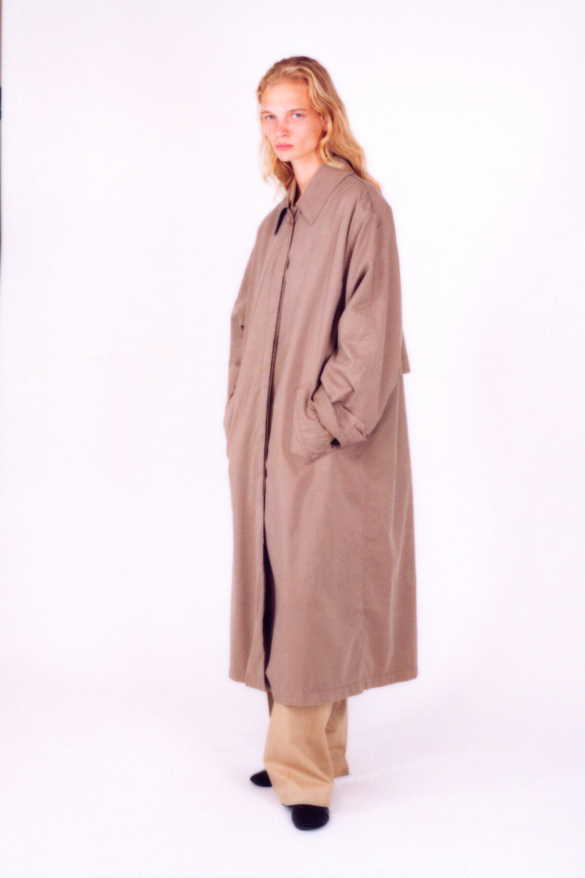 Full-body studio portrait of a woman wearing a long beige trench coat with hands in pockets, neutral backdrop, blonde hair.