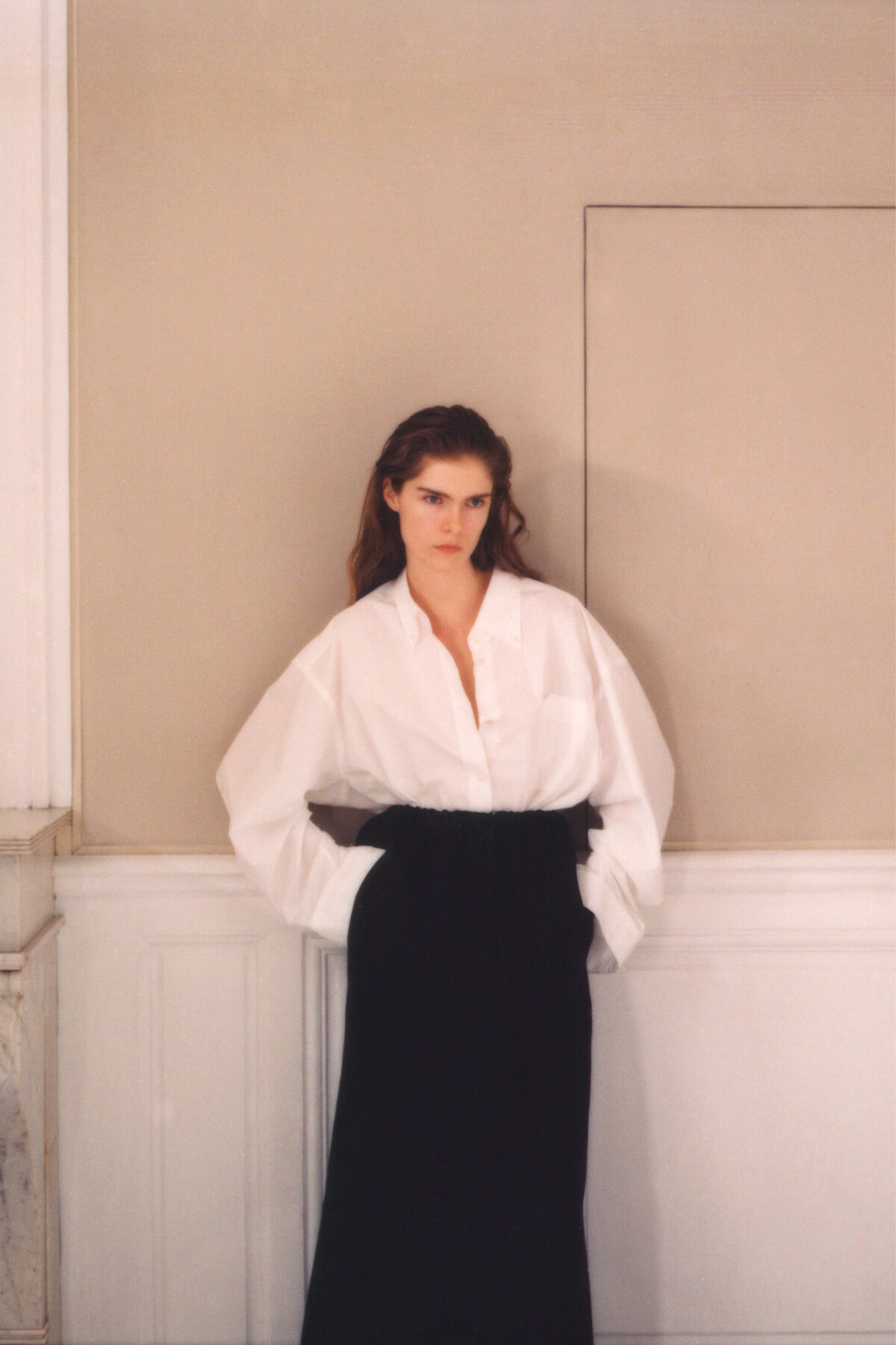 Young woman in a white blouse and long black skirt stands with her hands in pockets against a beige wall.