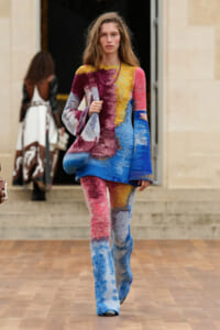 Model walks a wooden runway in a multicolor abstract-knit sweater and matching pants, carrying a slouchy bag.