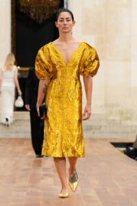 Model walks a runway wearing a crinkled metallic gold midi dress with dramatic puff sleeves and a deep V neckline.