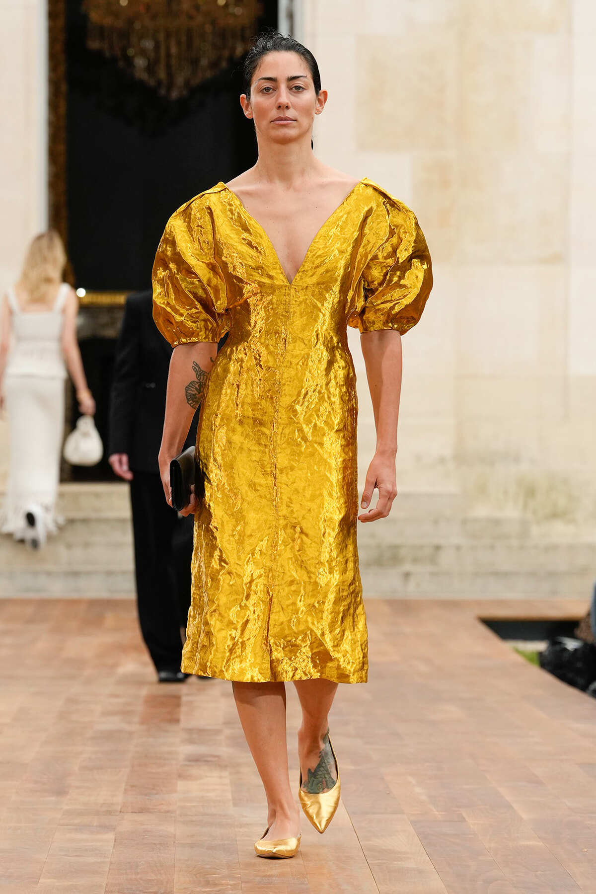 Model walks a runway wearing a crinkled metallic gold midi dress with dramatic puff sleeves and a deep V neckline.