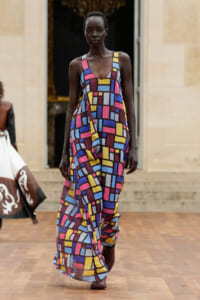 Black female model walks a runway in a sleeveless, floor-length maxi dress with a bright geometric color-block pattern.
