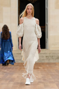 Model walking the runway in a cream crochet maxi dress with cold-shoulder sleeves and tasseled fringe hem, paired with white sneakers.