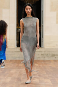 Model walking a runway in a silver, chainmail-inspired sleeveless dress with frayed hem and matching flats, during a fashion show.