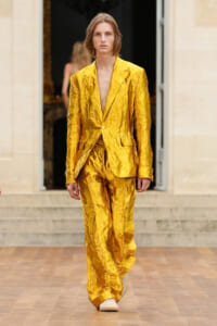 Model walks a runway in a crinkled, shiny gold suit with a single button blazer and wide-leg pants, beige sneakers visible at the bottom