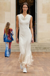 Model walking a fashion show runway in a sleeveless white textured maxi dress with a fringed hem and white sneakers.