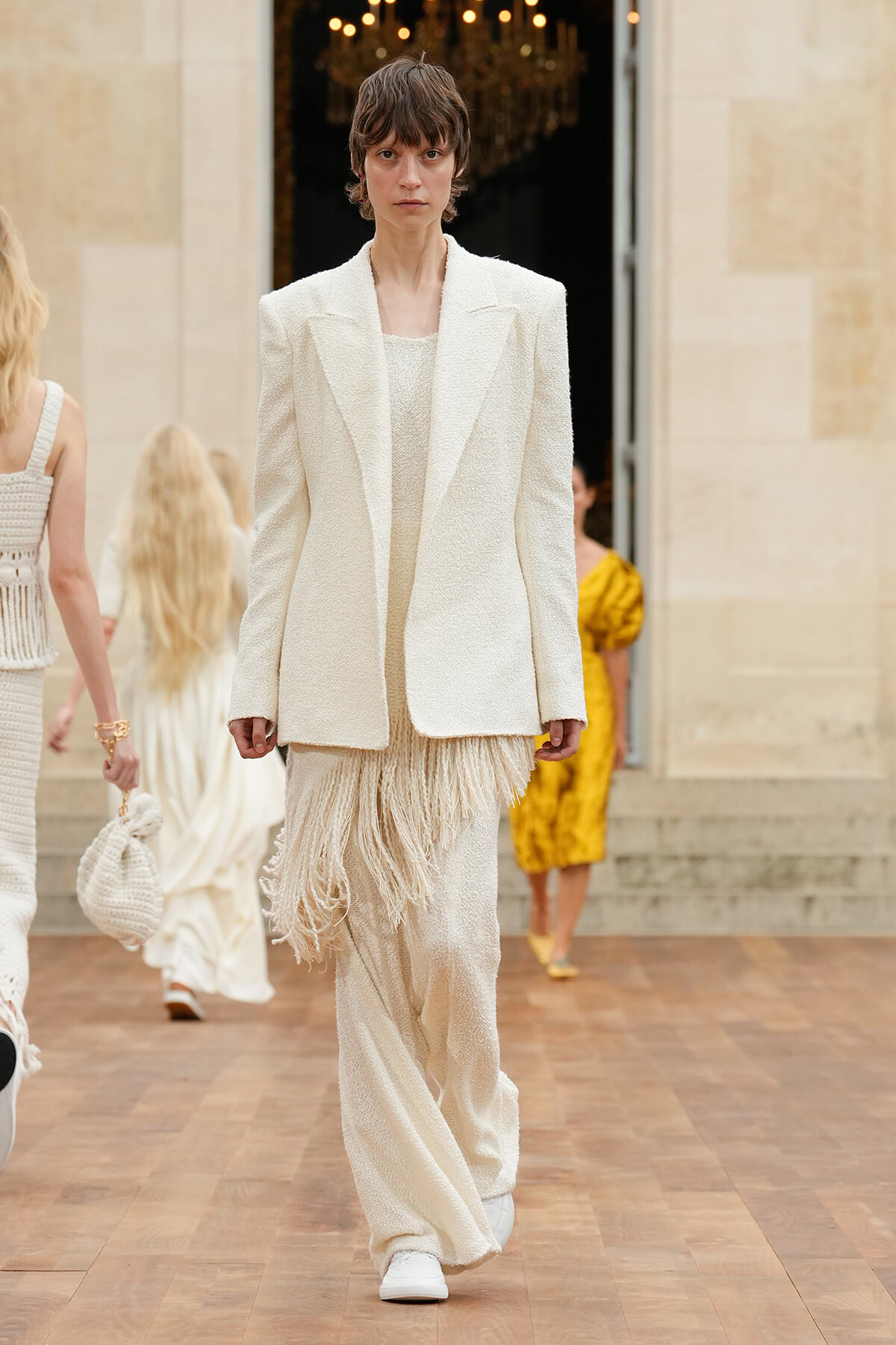 Model walks a fashion runway in a textured cream suit with a fringe-hem skirt and white sneakers.