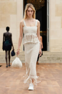 Model on a runway wearing a cream crochet fringe set and carrying a matching knit bag.