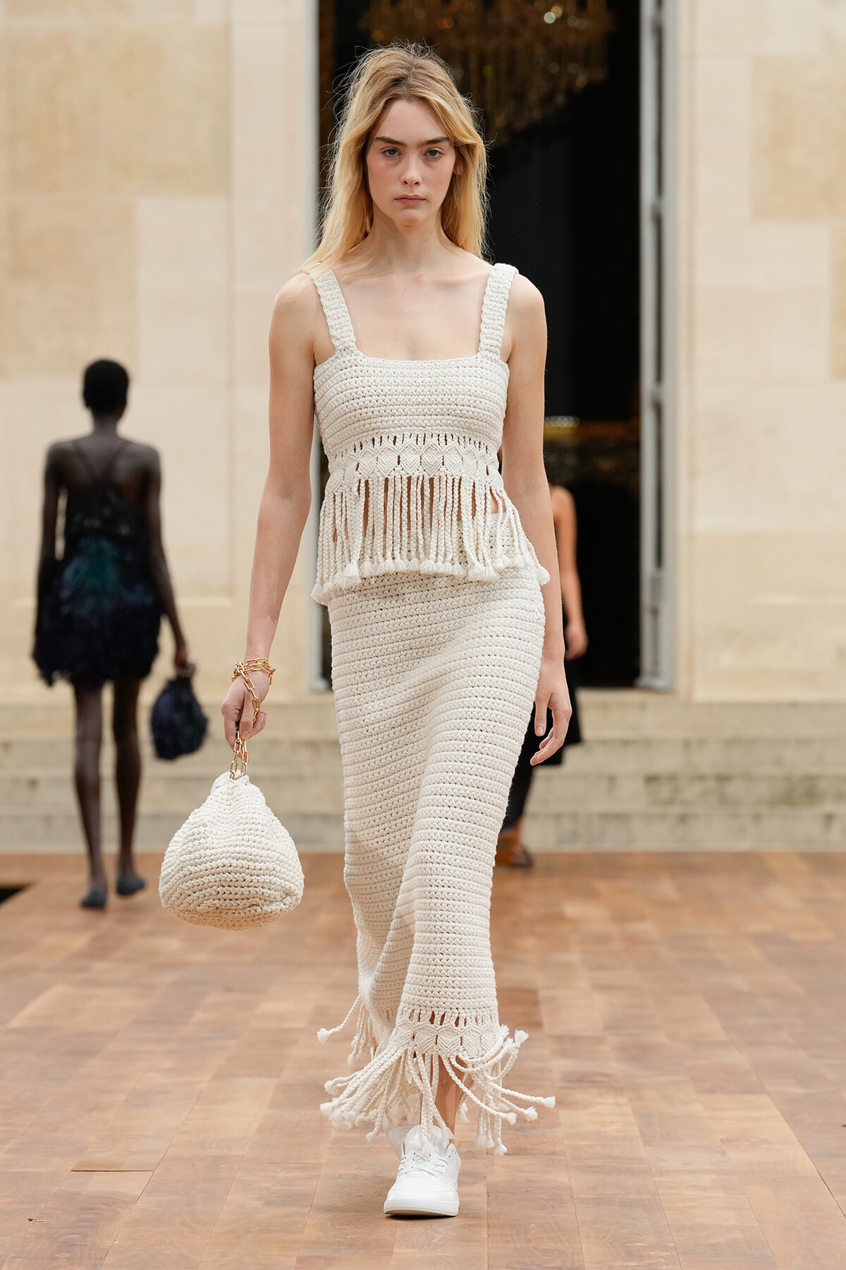 Model on a runway wearing a cream crochet fringe set and carrying a matching knit bag.