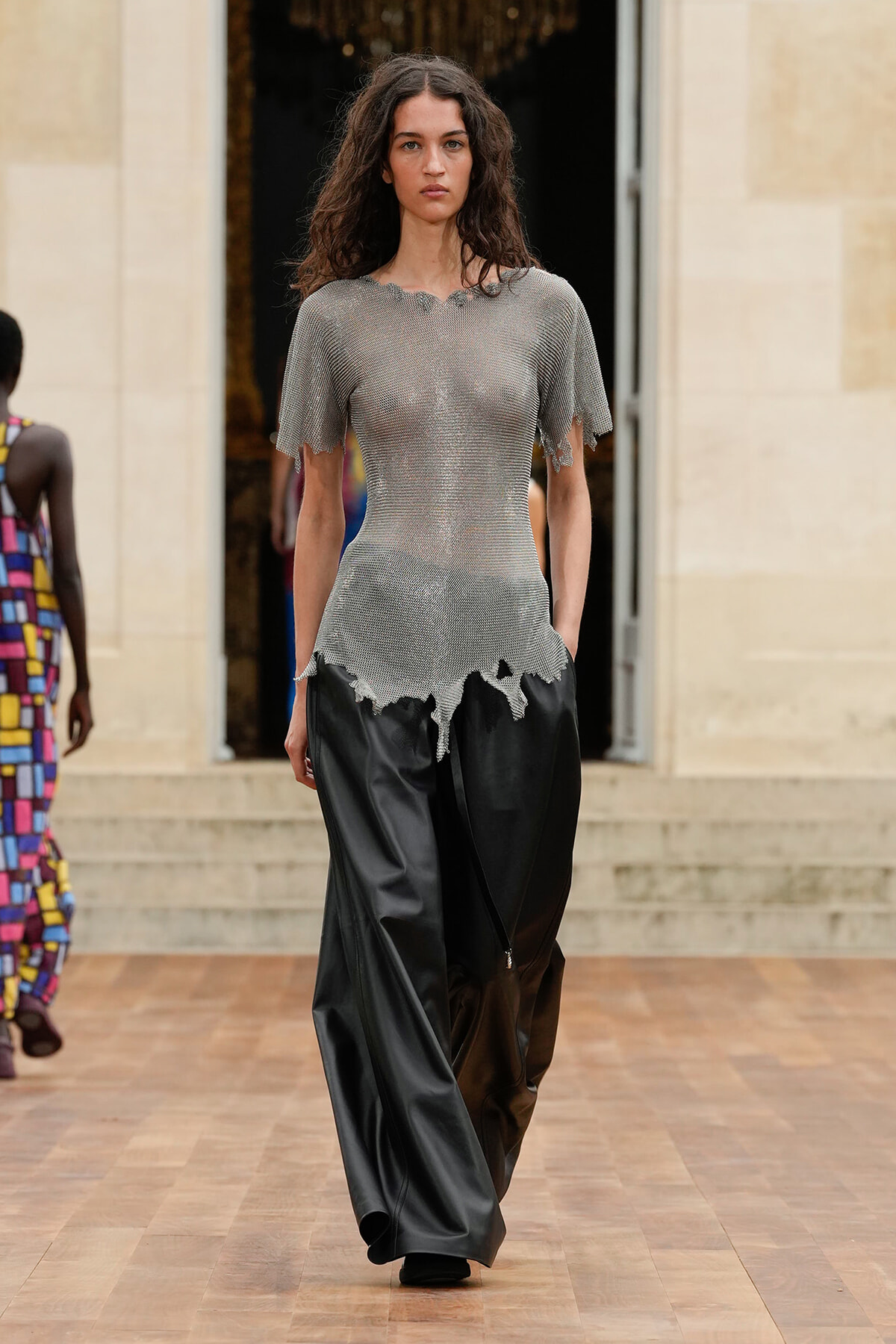 Model walking a fashion runway in a sheer silver knit top with a torn hem and wide black satin pants.