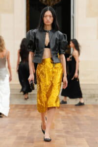 Model walks the runway in a black cropped leather jacket with puff sleeves, black bralette, and a crinkled gold midi skirt.