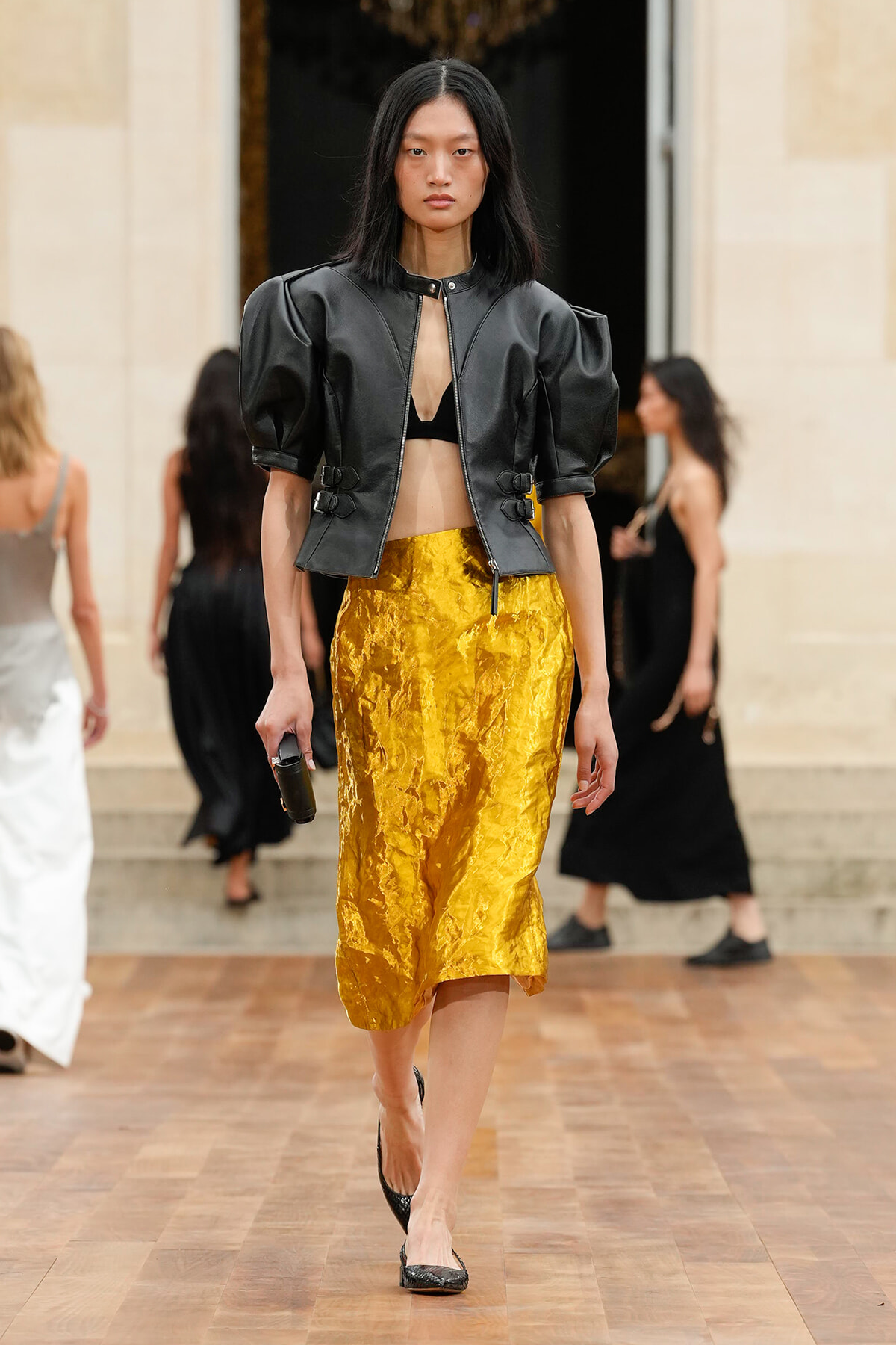 Model walks the runway in a black cropped leather jacket with puff sleeves, black bralette, and a crinkled gold midi skirt.