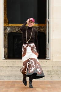 Woman standing on stone steps, tipping a pink cap, wearing a black turtleneck, a long brown-and-white patterned skirt, and black boots, with a chunky gold chain.