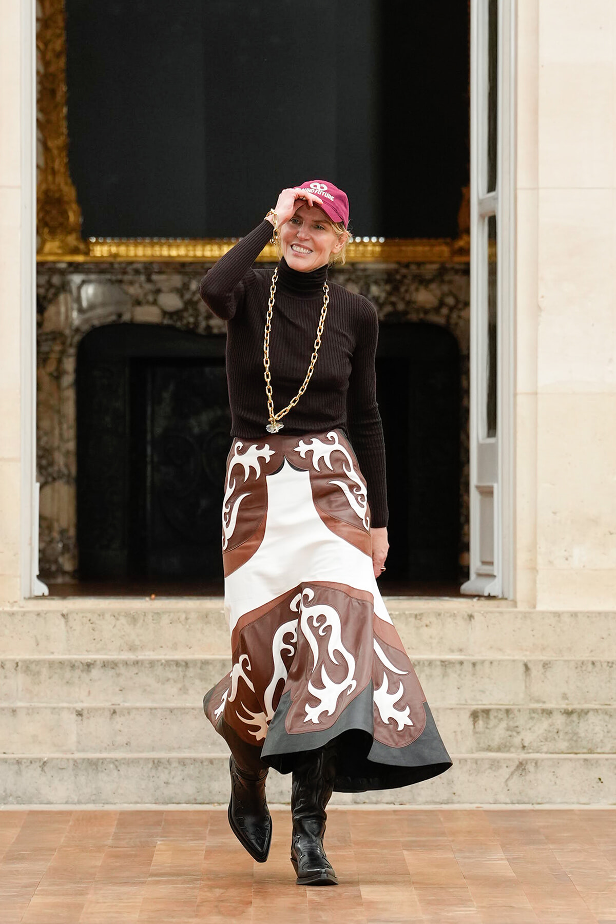 Woman standing on stone steps, tipping a pink cap, wearing a black turtleneck, a long brown-and-white patterned skirt, and black boots, with a chunky gold chain.
