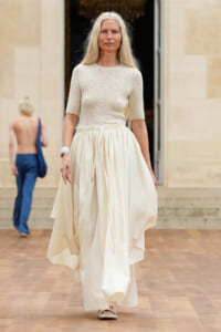 Blonde woman wearing a cream knit top and a long flowing ivory skirt walking on a wooden floor runway setting.