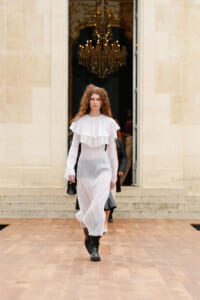 Model walks toward camera in a white, tiered ruffle dress with black boots, stepping on a wooden runway with a chandelier-lit doorway behind her.