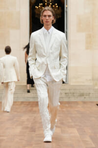 Male model in a cream textured suit walking down a runway, hands in pockets, white shirt and tie visible amid an elegant backdrop