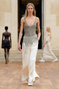 Model walking a runway in a sheer silver knit bodice and white satin wide-leg pants with fringe detailing at the hem.