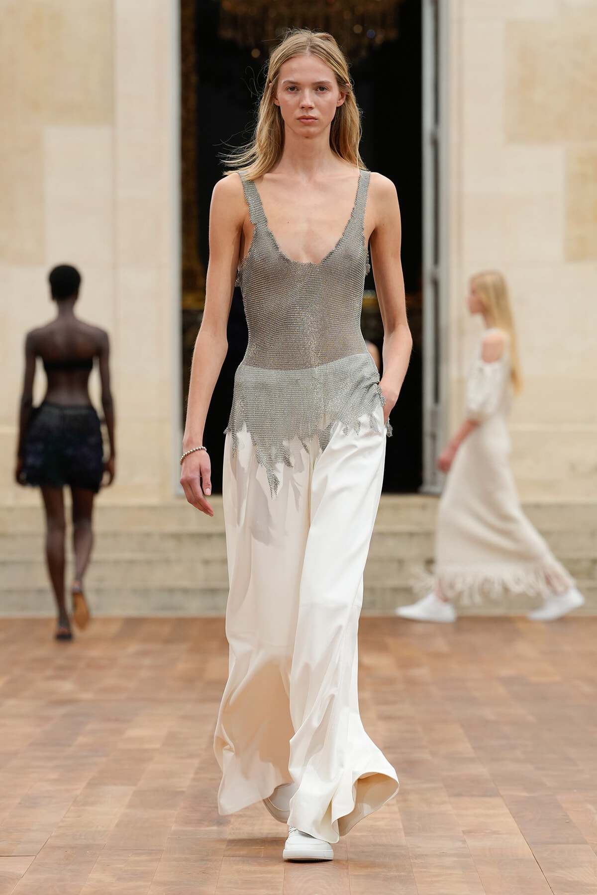 Model walking a runway in a sheer silver knit bodice and white satin wide-leg pants with fringe detailing at the hem.