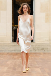Model walks a runway in a white satin slip dress with thin straps, gold necklace, and shiny gold heels outdoors.
