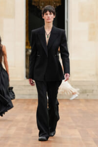 Model in a tailored black pantsuit with a plunging neckline and green pendant walks a runway, hands at sides.