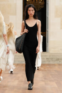 Model walking on a runway in a black sleeveless jumpsuit, carrying a large black handbag over her shoulder.