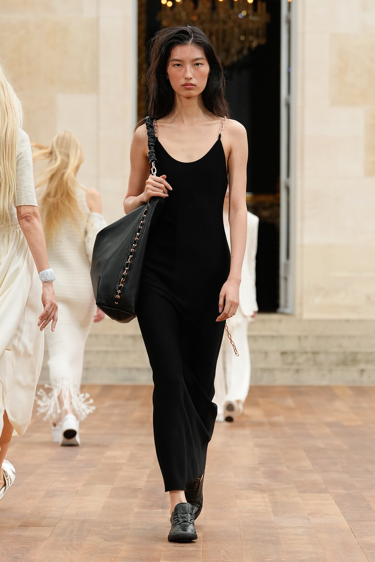 Model walking on a runway in a black sleeveless jumpsuit, carrying a large black handbag over her shoulder.