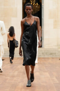 Model walking a runway in a black satin slip dress with thin straps and a silver necklace.
