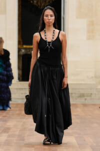 Model walking in a black tank top and full black skirt, chunky necklace, carrying a black bag indoors.