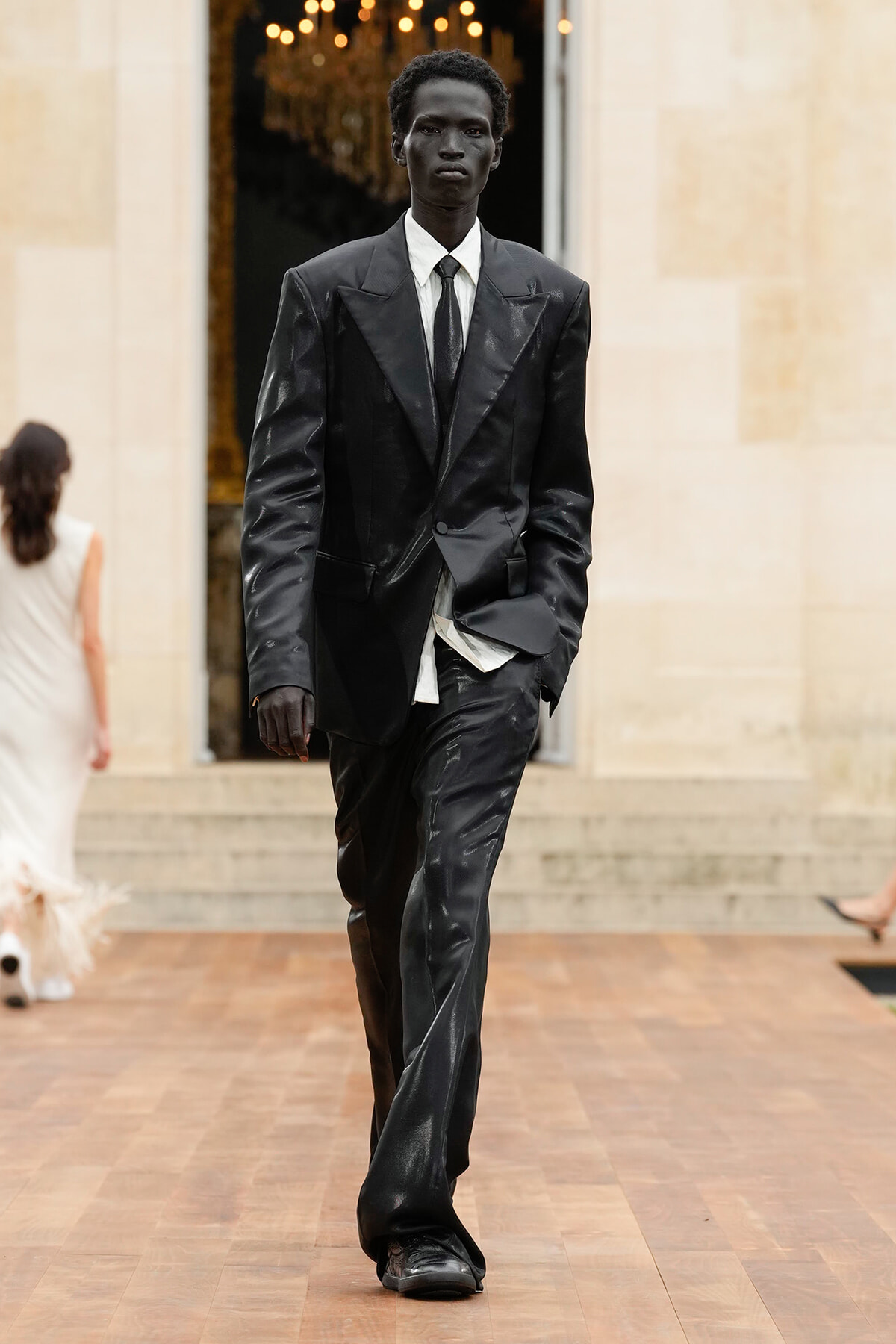 Male model in a black oversized suit and white shirt walks down a runway, hands in pockets, in a fashion show setting with warm lighting.
