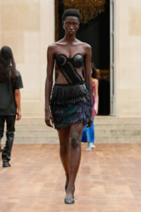 Model walking a runway in a black sculptural corset top and a multi-layered feathered mini skirt studded with metal accents, during a fashion show.