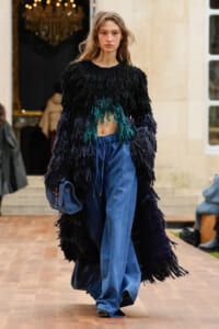 Model on a runway wearing a black shaggy fringed coat with teal fringe, loose blue denim pants, and a blue handbag.