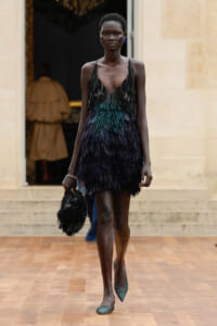 Model walking a runway in a black-to-teal feathered fringe mini dress, matching fringe bag in hand, blue earrings and green pointed heels.