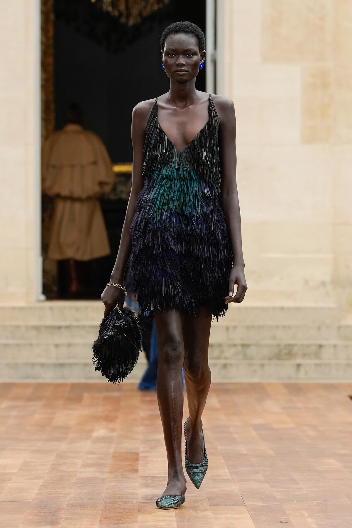 Model walking a runway in a black-to-teal feathered fringe mini dress, matching fringe bag in hand, blue earrings and green pointed heels.